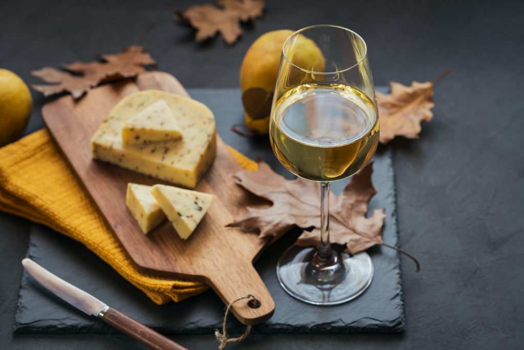 A glass of white wine served with cheese in a cutting board on dark background